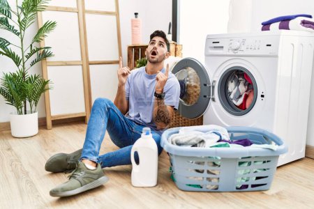 Téléchargez les photos : Young hispanic man putting dirty laundry into washing machine amazed and surprised looking up and pointing with fingers and raised arms. - en image libre de droit