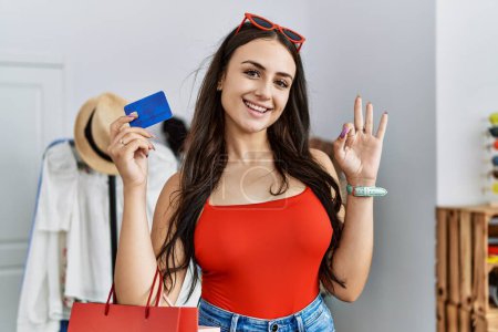 Foto de Joven morena sosteniendo bolsas de compras y tarjeta de crédito haciendo signo bien con los dedos, sonriendo gesto amistoso excelente símbolo - Imagen libre de derechos