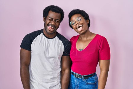Foto de Joven pareja afroamericana de pie sobre fondo rosa sacando la lengua feliz con expresión divertida. concepto de emoción. - Imagen libre de derechos