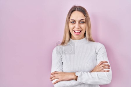 Foto de Mujer rubia joven con suéter blanco sobre fondo rosa cara feliz sonriendo con los brazos cruzados mirando a la cámara. persona positiva. - Imagen libre de derechos