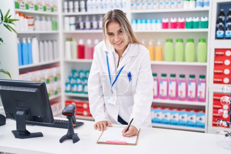 Téléchargez les photos : Young blonde woman pharmacist writing on document at pharmacy - en image libre de droit