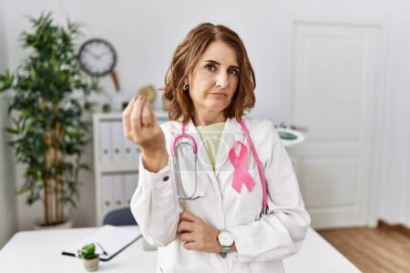 Téléchargez les photos : Médecin d'âge moyen femme portant un ruban de cancer rose sur l'uniforme faisant geste italien avec la main et les doigts expression confiante - en image libre de droit