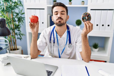 Téléchargez les photos : Young hispanic dietitian man holding doughnut and apple puffing cheeks with funny face. mouth inflated with air, catching air. - en image libre de droit