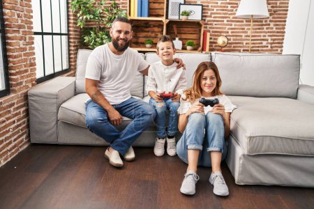 Foto de Family of three playing video game sitting on the sofa looking positive and happy standing and smiling with a confident smile showing teeth - Imagen libre de derechos