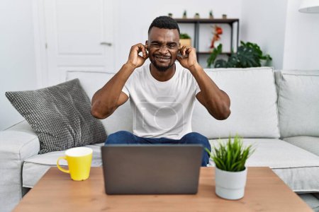 Foto de Young african man using laptop at home covering ears with fingers with annoyed expression for the noise of loud music. deaf concept. - Imagen libre de derechos