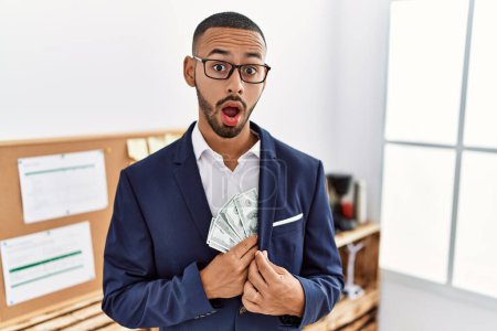 Téléchargez les photos : Jeune homme afro-américain cachant des dollars en veste dans le visage de choc, l'air sceptique et sarcastique, surpris avec la bouche ouverte - en image libre de droit