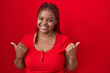 Foto de Mujer afroamericana con el pelo trenzado de pie sobre el signo de éxito de fondo rojo haciendo gesto positivo con la mano, pulgares hacia arriba sonriendo y feliz. expresión alegre y gesto ganador. - Imagen libre de derechos