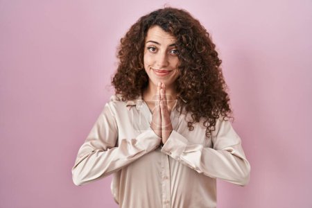 Téléchargez les photos : Hispanic woman with curly hair standing over pink background praying with hands together asking for forgiveness smiling confident. - en image libre de droit