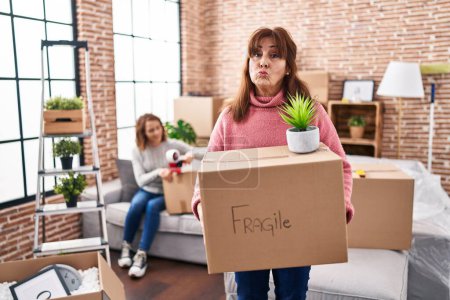 Photo for Mother and daughter moving to a new home holding cardboard box looking at the camera blowing a kiss being lovely and sexy. love expression. - Royalty Free Image