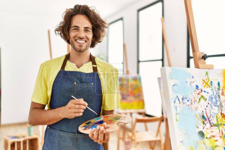 Photo for Young hispanic artist man smiling happy holding paintbrush and palette at art studio. - Royalty Free Image