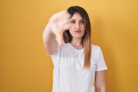 Foto de Young brunette woman standing over yellow background looking unhappy and angry showing rejection and negative with thumbs down gesture. bad expression. - Imagen libre de derechos
