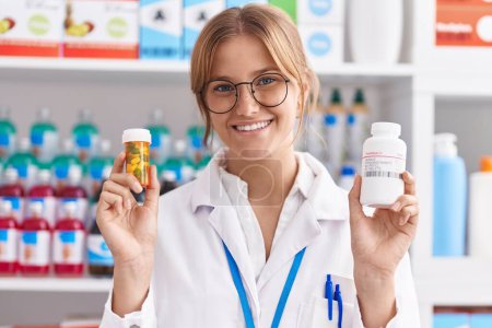 Téléchargez les photos : Jeune fille blonde pharmacien souriant confiant tenant des bouteilles de pilules à la pharmacie - en image libre de droit
