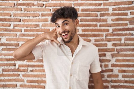 Téléchargez les photos : Homme arabe avec barbe debout sur le mur de briques arrière-plan souriant faire geste de téléphone avec la main et les doigts comme parler au téléphone. communiquer les concepts. - en image libre de droit