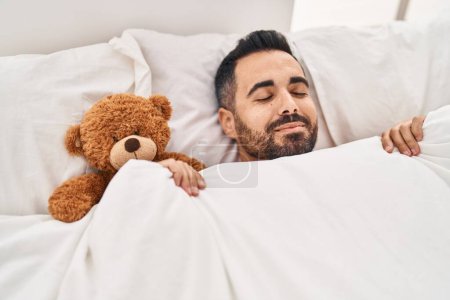 Joven hispano acostado en la cama durmiendo con un osito de peluche en el dormitorio