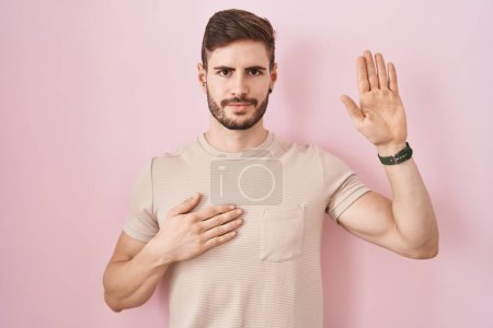 Téléchargez les photos : Homme hispanique avec la barbe debout sur fond rose jurant avec la main sur la poitrine et la paume ouverte, faisant un serment de fidélité promesse - en image libre de droit