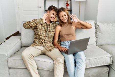 Photo for Young hispanic couple having video call using laptop sitting on the sofa at home. - Royalty Free Image