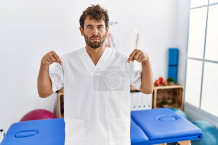 Joven hombre guapo fisioterapeuta que trabaja en la clínica de recuperación del dolor apuntando hacia abajo mirando triste y molesto, indicando la dirección con los dedos, infeliz y deprimido. 