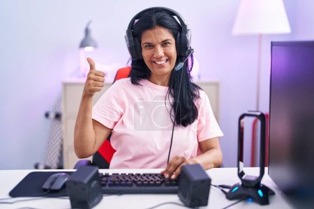 Foto de Mature hispanic woman playing video games at home doing happy thumbs up gesture with hand. approving expression looking at the camera showing success. - Imagen libre de derechos