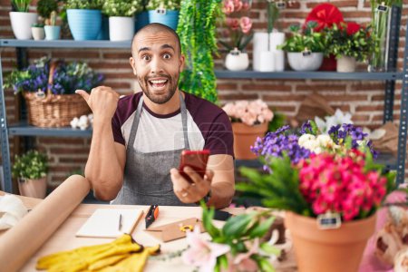 Photo for Hispanic man with beard working at florist shop with smartphone pointing thumb up to the side smiling happy with open mouth - Royalty Free Image