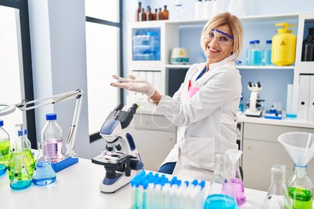 Téléchargez les photos : Moyen Age femme blonde travaillant au laboratoire scientifique pointant de côté avec les mains ouvertes paumes montrant l'espace de copie, présentant la publicité souriant excité heureux - en image libre de droit