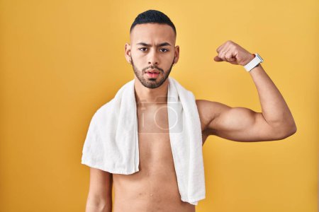 Foto de Joven hombre hispano de pie sin camisa con toalla fuerte persona mostrando el músculo del brazo, confiado y orgulloso de poder - Imagen libre de derechos