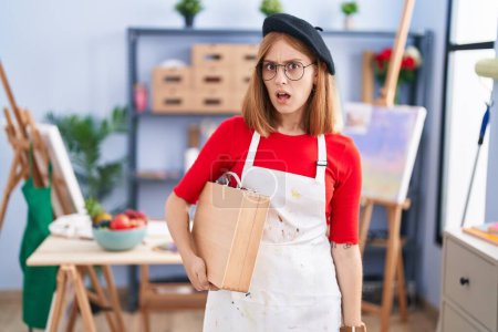 Photo for Young redhead woman at art studio holding art case in shock face, looking skeptical and sarcastic, surprised with open mouth - Royalty Free Image