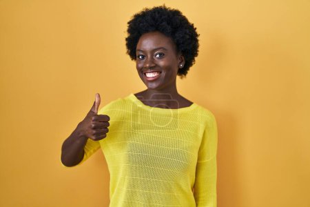 Téléchargez les photos : Jeune femme africaine debout sur le studio jaune faisant des pouces heureux geste avec la main. approuvant l'expression regardant la caméra montrant le succès. - en image libre de droit