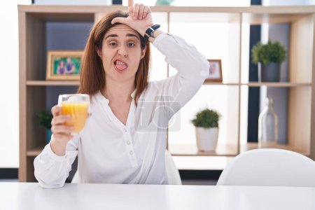 Photo for Brunette woman drinking glass of orange juice making fun of people with fingers on forehead doing loser gesture mocking and insulting. - Royalty Free Image