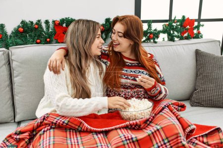 Téléchargez les photos : Femme couple étreignant l'autre regarder la télévision assis par décor de Noël à la maison - en image libre de droit