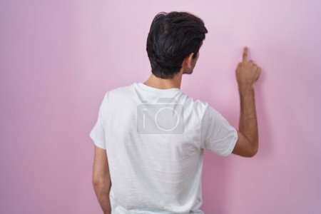 Foto de Young hispanic man standing over pink background posing backwards pointing ahead with finger hand - Imagen libre de derechos