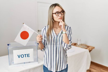 Foto de Asian young woman at political campaign election holding japan flag serious face thinking about question with hand on chin, thoughtful about confusing idea - Imagen libre de derechos