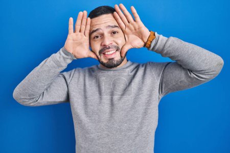 Photo for Hispanic man standing over blue background smiling cheerful playing peek a boo with hands showing face. surprised and exited - Royalty Free Image