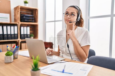 Young hispanic woman wearing call center agent headset at the office serious face thinking about question with hand on chin, thoughtful about confusing idea 