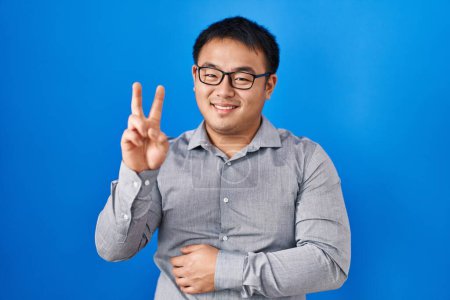 Foto de Young chinese man standing over blue background smiling with happy face winking at the camera doing victory sign. number two. - Imagen libre de derechos