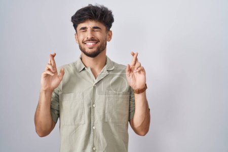 Téléchargez les photos : Homme arabe avec barbe debout sur fond blanc geste doigt croisé souriant avec espoir et les yeux fermés. chance et concept superstitieux. - en image libre de droit