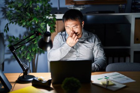 Photo for Young chinese man working using computer laptop at night smelling something stinky and disgusting, intolerable smell, holding breath with fingers on nose. bad smell - Royalty Free Image