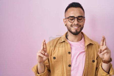 Téléchargez les photos : Jeune homme hispanique debout sur fond rose geste doigt croisé souriant avec espoir et les yeux fermés. chance et concept superstitieux. - en image libre de droit