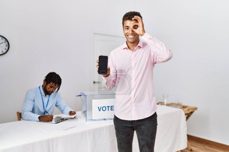 Téléchargez les photos : Les jeunes hommes hispaniques à l'élection de campagne politique tenant smartphone souriant heureux faire ok signe avec la main sur les yeux regardant à travers les doigts - en image libre de droit