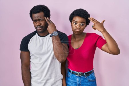 Foto de Joven pareja afroamericana de pie sobre fondo rosa disparando y matándose señalando la mano y los dedos a la cabeza como arma, gesto suicida. - Imagen libre de derechos