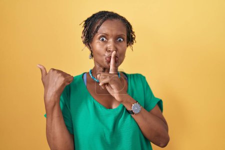 Téléchargez les photos : Femme africaine avec dreadlocks debout sur fond jaune demandant à être calme avec le doigt sur les lèvres pointant avec la main sur le côté. silence et concept secret. - en image libre de droit