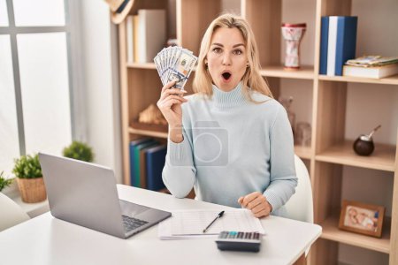 Photo for Young caucasian woman using laptop holding dollars banknotes scared and amazed with open mouth for surprise, disbelief face - Royalty Free Image