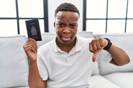 Téléchargez les photos : Jeune homme africain tenant un passeport canada avec le visage en colère, signe négatif montrant aversion avec les pouces vers le bas, concept de rejet - en image libre de droit