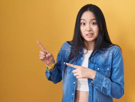 Photo for Young asian woman standing over yellow background pointing aside worried and nervous with both hands, concerned and surprised expression - Royalty Free Image