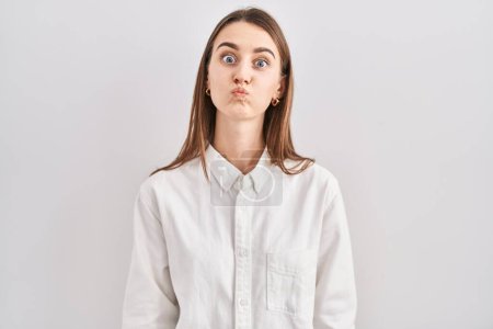 Foto de Young caucasian woman standing over isolated background puffing cheeks with funny face. mouth inflated with air, crazy expression. - Imagen libre de derechos