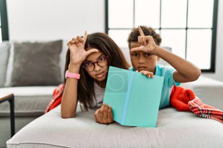 Photo for Two siblings lying on the sofa reading a book making fun of people with fingers on forehead doing loser gesture mocking and insulting. - Royalty Free Image