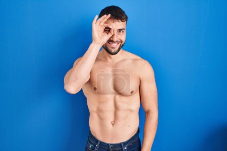 Photo for Handsome hispanic man standing shirtless doing ok gesture with hand smiling, eye looking through fingers with happy face. - Royalty Free Image