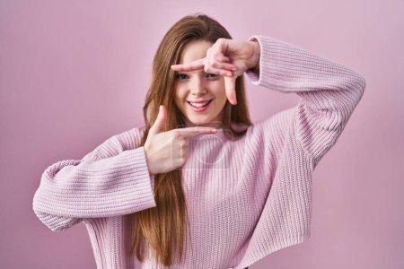 Foto de Mujer caucásica joven de pie sobre fondo rosa sonriendo haciendo marco con las manos y los dedos con la cara feliz. concepto de creatividad y fotografía. - Imagen libre de derechos