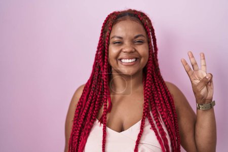 Téléchargez les photos : Femme afro-américaine avec les cheveux tressés debout sur fond rose montrant et pointant vers le haut avec les doigts numéro trois tout en souriant confiant et heureux. - en image libre de droit