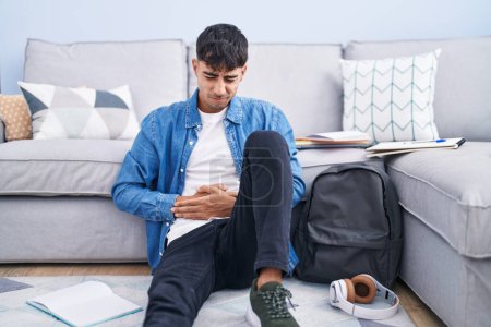 Foto de Young hispanic man sitting on the floor studying for university with hand on stomach because indigestion, painful illness feeling unwell. ache concept. - Imagen libre de derechos