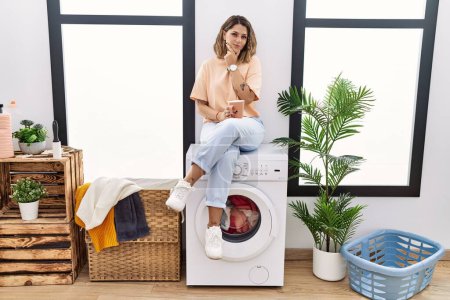 Foto de Young hispanic woman drinking coffee waiting for washing machine at laundry room looking confident at the camera smiling with crossed arms and hand raised on chin. thinking positive. - Imagen libre de derechos